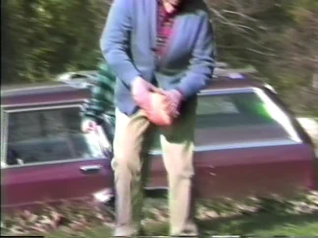 A young boy in a plaid shirt practices kicking a football held by his father on a lawn next to parks and the father crouches to steady the ball before the boy kicks it and runs toward a brick house in a wooded suburban yard