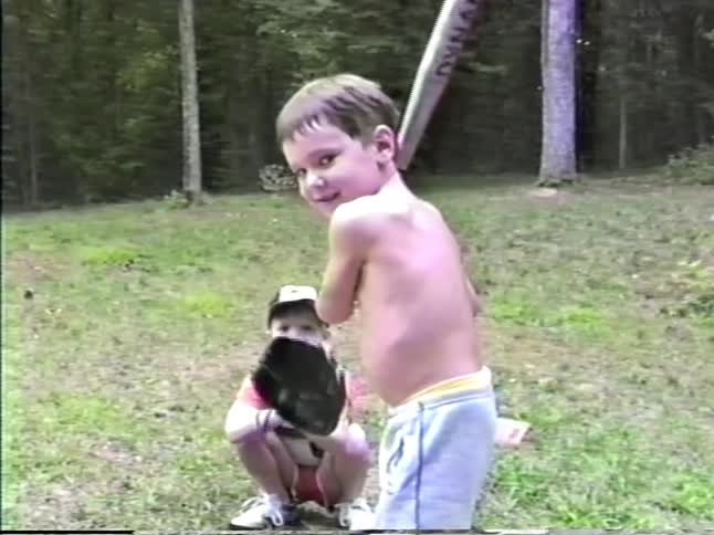A young boy with no shirt and blue shorts swings a silver baseball bat while another boy wearing a baseball cap and glove crouches behind him in a grassy yard near a forest