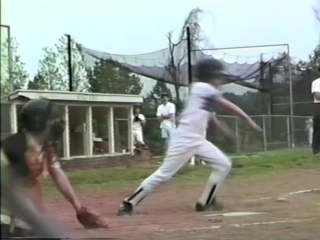 A young baseball player in a white uniform hits a pitch and sprints toward first base on a dirt field while a catcher watches from behind home plate near a wooden dugout and a chainlink fence