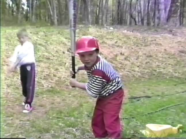 Boys practice swinging and hitting a baseball while on the lawn of their rural home on a summer day