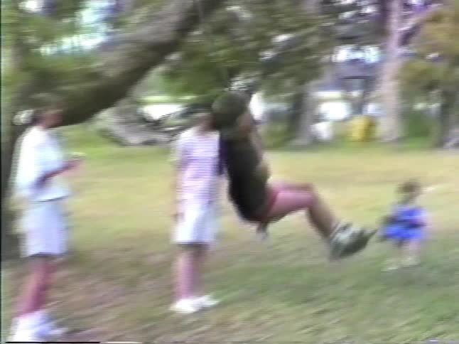 A young boy in a black tshirt and pink shorts laughs while swinging on a rope tied to a large tree in a grassy backyard while watched by other people near by