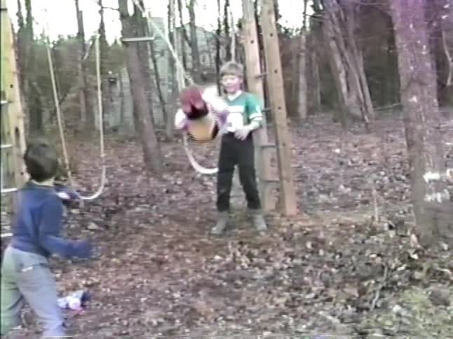 Some children play on a wooden climbing toy and swings in a wooded backyard filled with fallen leaves and trees