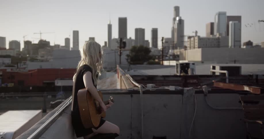 A woman leans against a roof top and plays guitar