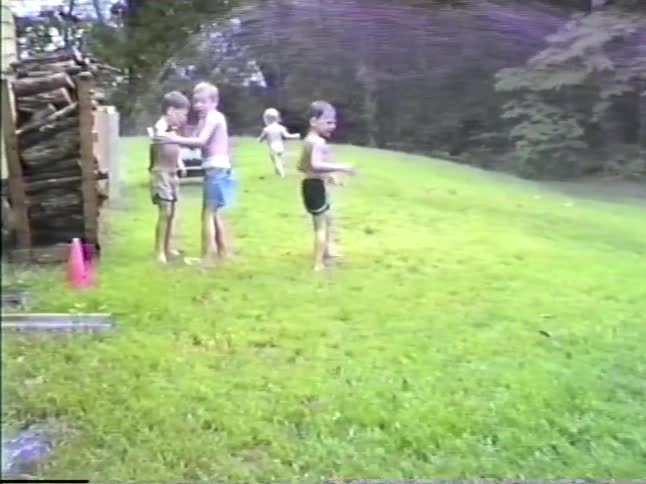 A group of young children without shirts run and play in a grassy yard while being sprayed with water from a garden hose held by a man sitting in a lawn chair