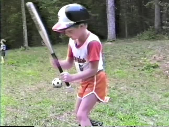 Child holding a bat in a grassy area while other children play in the background