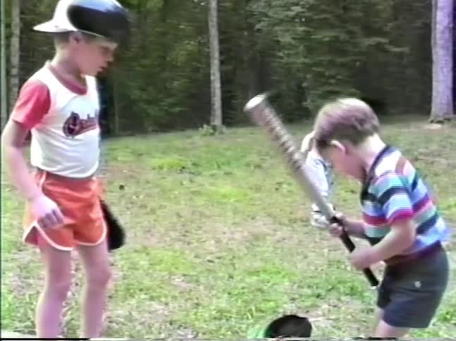 Children playing baseball on a grassy area near a wooded area
