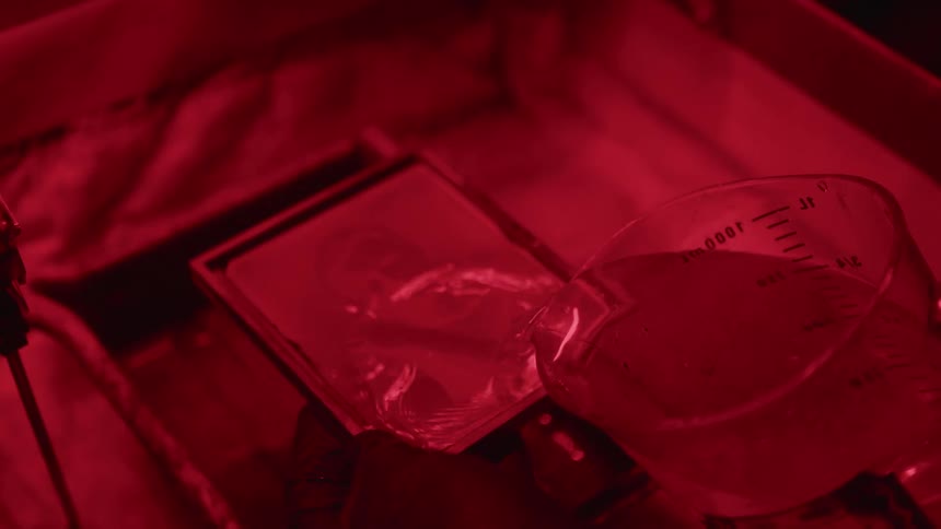 Photographer pouring solution into a container developing a picture or a man under a red light in the darkroom