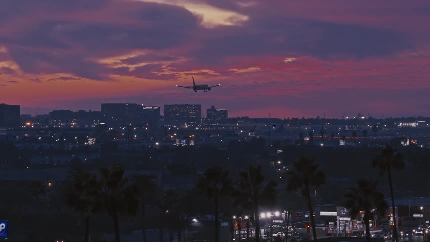 Aerial view of a city skyline with an airplane descending above silhouetted palm trees under a sunset sky