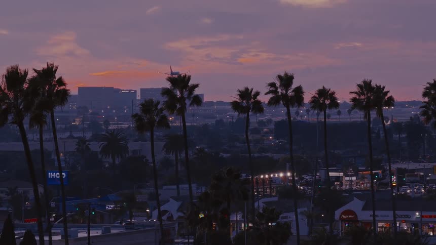 A commercial airplane descends toward a runway over a silhouetted row of palm trees and a suburban commercial district during a purple and orange sunset