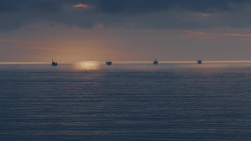Four offshore oil platforms sit silhouetted against a hazy sunset on a calm ocean under a dark cloudy sky as light reflects off the waters surface toward the horizon