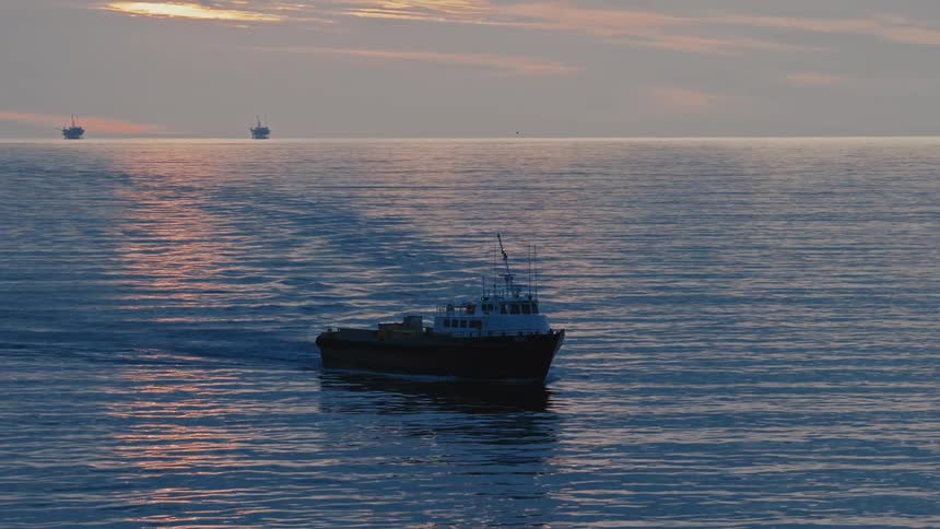 A small workboat moves steadily across a calm reflective sea at sunset with several distant offshore oil platforms silhouetted along the horizon beneath a softly glowing cloudstreaked sky