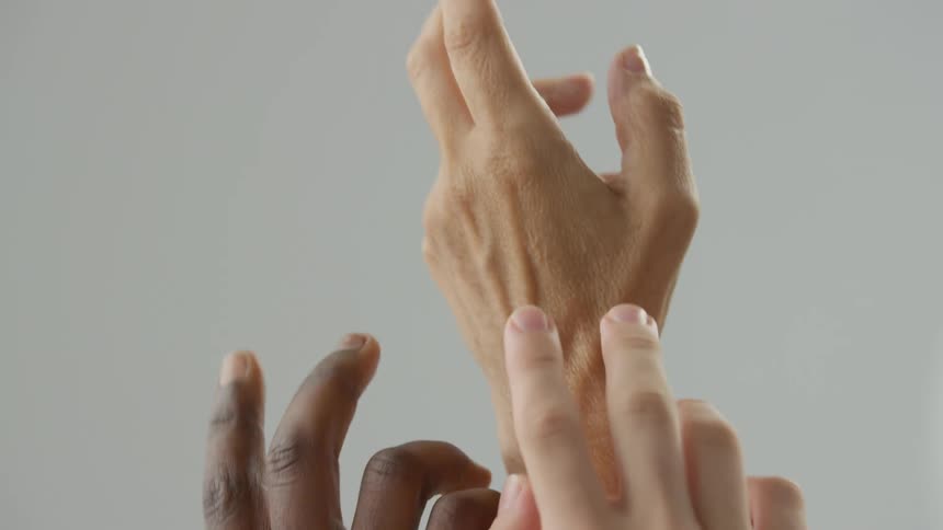 Multiple hands with different skin tones reach upward and gently touch each others fingers and wrists against a neutral grey background in a studio setting