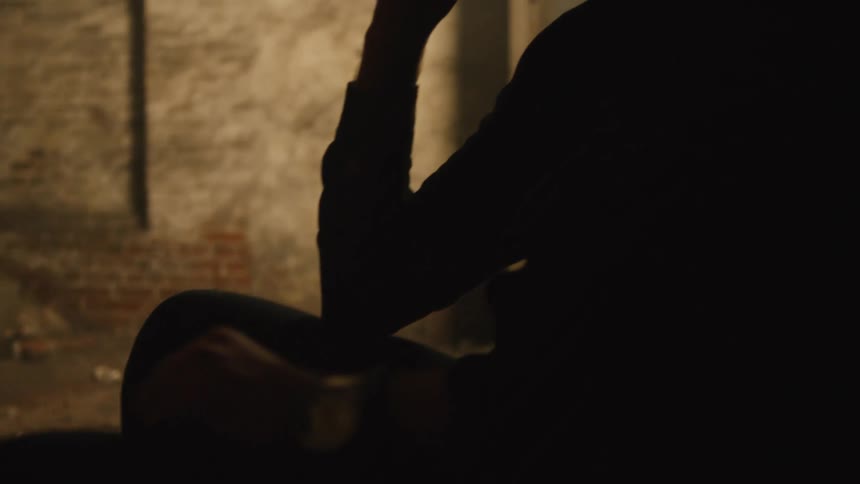A person sits in a dimly lit industriallooking space with their silhouette cast against a rough brick wall the camera pans up to reveal a man with a prominent beard and a hat looking off into the distance