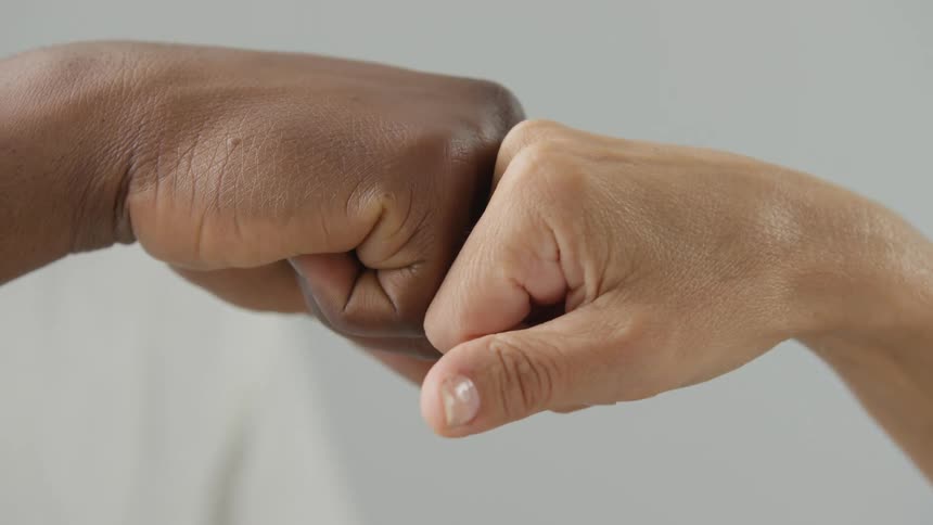 Closeup of two people of different ethnicities performing a fist bump with their hands against a plain gray background