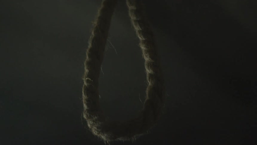 A thick textured rope is tied into a hangmans noose and suspended in a dimly lit environmentthe camera slowly tilts upward along the rope emphasizing its coarse fibers against the dark shadowy background