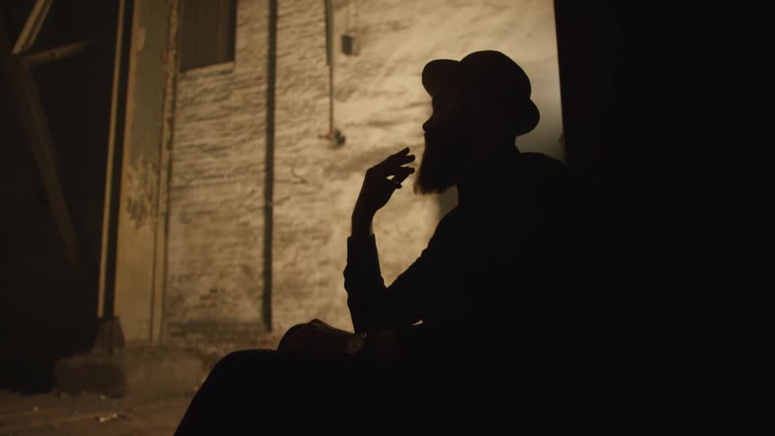 Silhouetted man strokes his beard while sitting against a wall