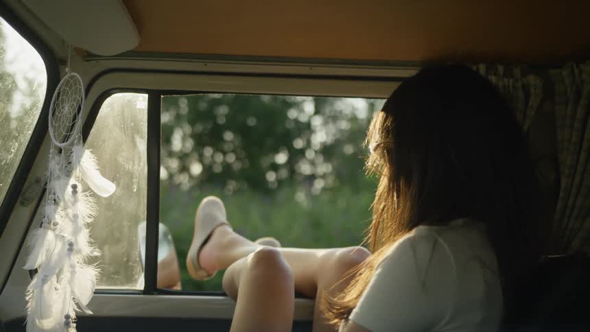 A woman sits comfortably inside a vintage van resting her feet on the open window frame while looking out at a sundrenched fieldthe scene captures a peaceful bohemian atmosphere emphasized by a white dreamcatcher hanging near the glass and the soft