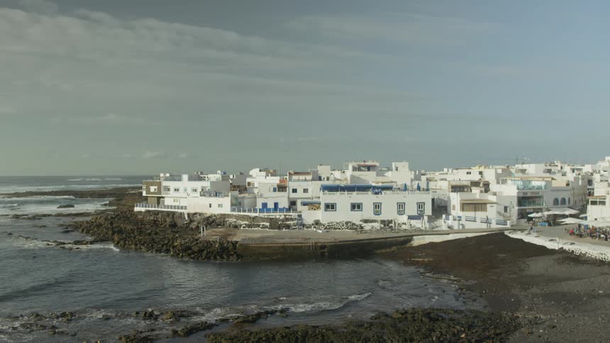 White coastal buildings cluster on a rocky shoreline beside the ocean under an overcast sky