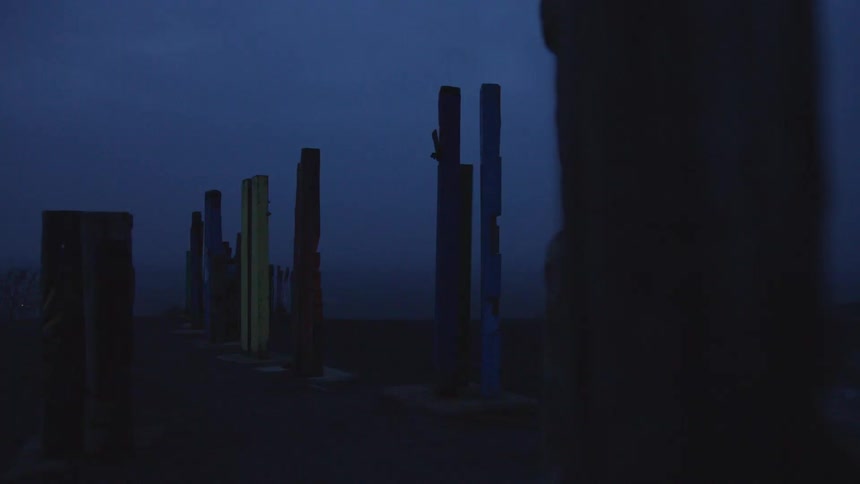 A series of tall narrow rectangular pillars of varying colors stand in two parallel rows outdoors at duskthe camera slowly pans to the right showing these structures positioned along a walkway against a dark foggy landscape