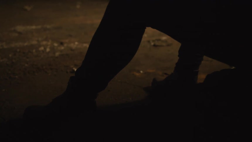 In a lowangle dimly lit shot the silhouette of a person wearing dark boots and trousers is seen crouched or sitting on a gritty debrisstrewn grounda faint light from above casts a warm glow on the textured earth while the figure remains largely i