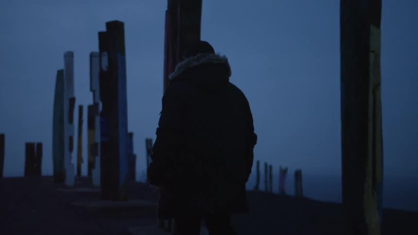A person walks along a path lined with tall wooden pillars under a dark blue twilight sky