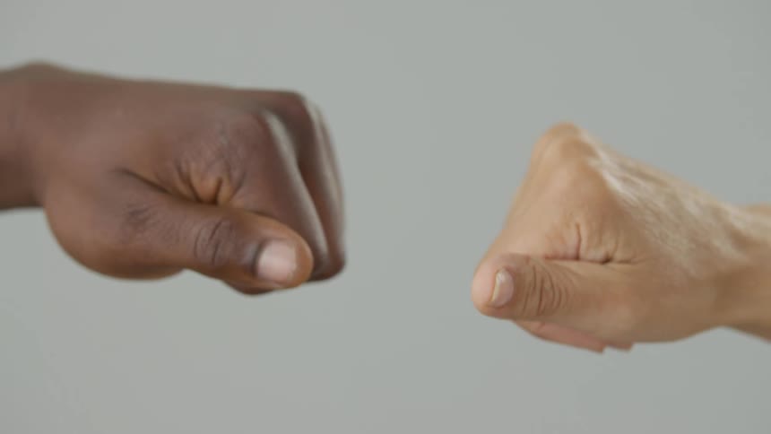 Closeup of two people of different ethnicities performing a fist bump with their hands against a plain gray background