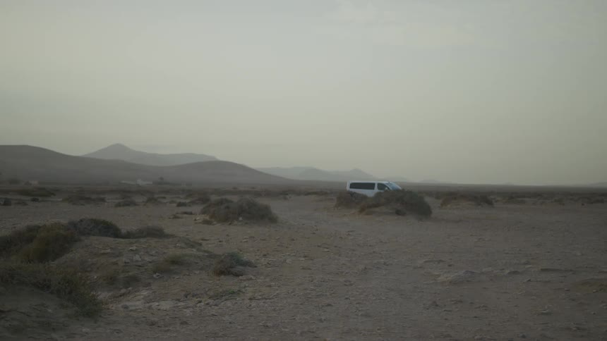 A white van drives across a vast dusty desert landscape under a hazy sky the vehicle moves steadily through the barren terrain passing small shrubs and distant hills