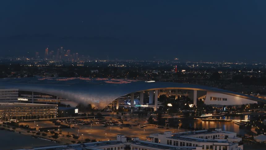 The footage shows an aerial evening view of sofi stadium in inglewood featuring its illuminated sweeping canopy and the surrounding parking areasin the distant background the glowing skyline of downtown los angeles is visible under a dark twilight s
