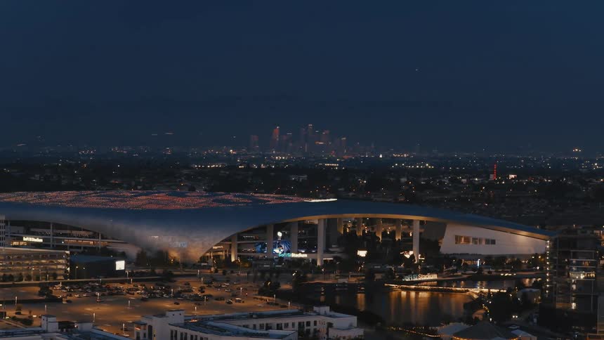 This video features a highangle night shot of sofi stadium in inglewood california with its distinctive translucent canopy illuminatedin the background the downtown los angeles skyline glows against the dark twilight sky