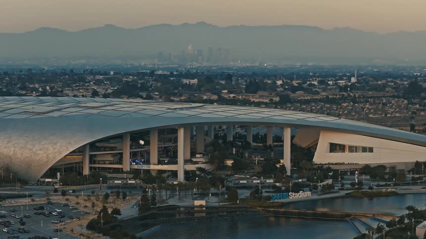 A massive modern stadium with a sweeping metallic roof dominating an urban landscape surrounded by roads parked cars and water features with a hazy city skyline and mountains receding into the background