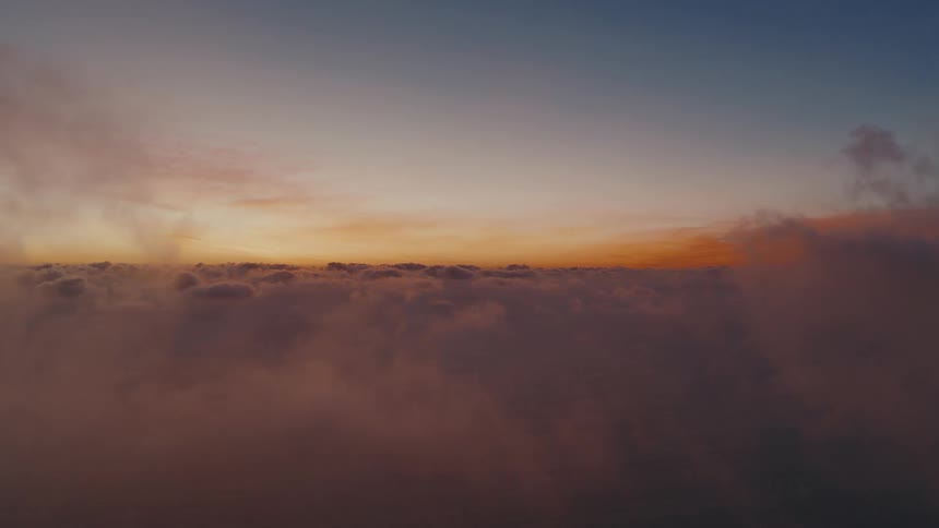 The camera glides over a dense blanket of clouds during sunset revealing a glowing orange horizon that fades into a deep blue twilight sky while wisps of mist pass through the foreground