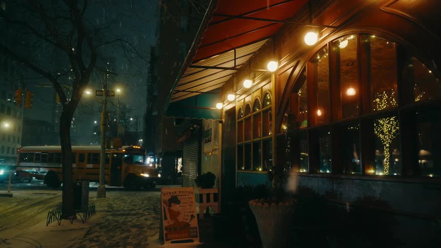 A nighttime street scene with snow falling as a bus turns right at an intersection