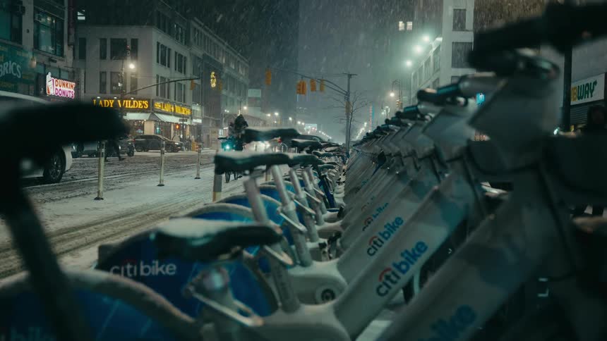 A person wearing a black hooded jacket with a yellow strap rides a bicycle down a snowy city street at night past a row of parked bicycles