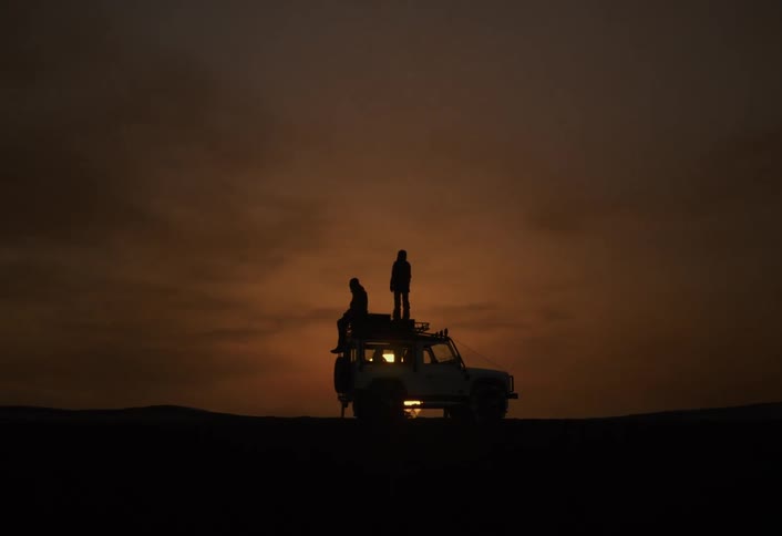 Silhouette of two people on top of a vehicle in the desert with the sun in the background