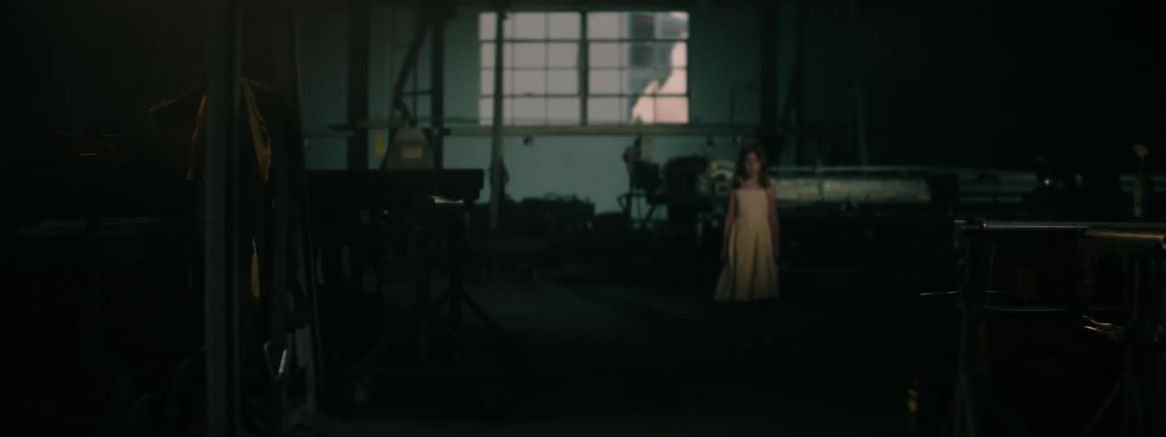 A young girl walks through a dim industrial workshop surrounded by machinery