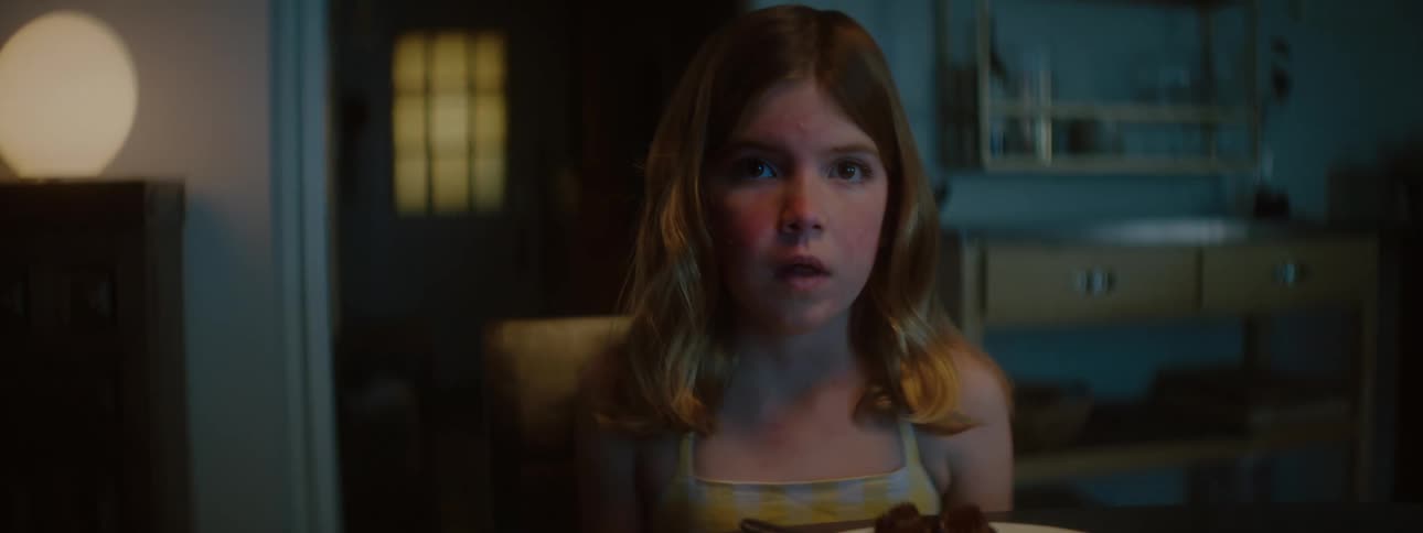 A young girl sits at a table staring forward with a frightened expression in a dim room