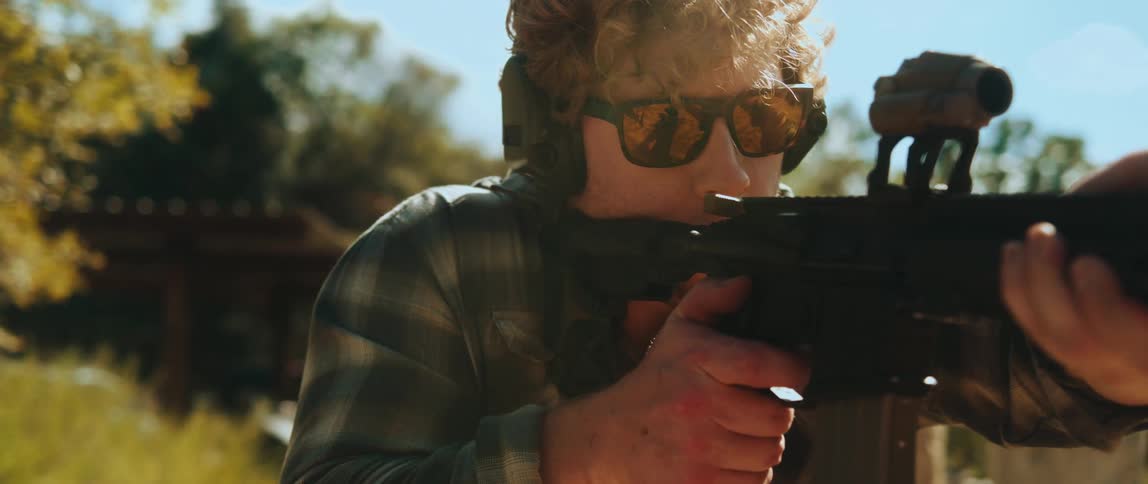 A man with curly hair wears sunglasses and ear protection while aiming a rifle toward the camerathe camera pans around to his side as he maintains his focus and grip on the firearm in an outdoor setting
