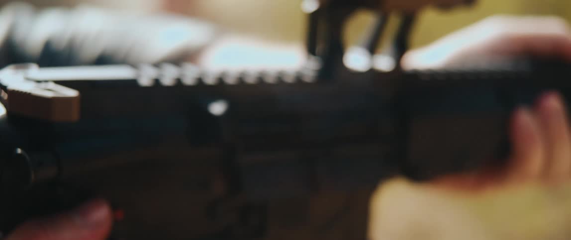 A rifle firing as a spent casing ejects from the chamber midair with the firearms rail and barrel partially in focus and a hand gripping it against a blurred background