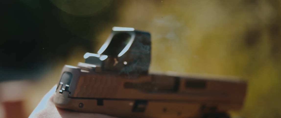 A handgun fires in slow motion showing the slide cycling back to eject a spent brass casing while a small puff of smoke and muzzle flash emerge against a blurred outdoor background