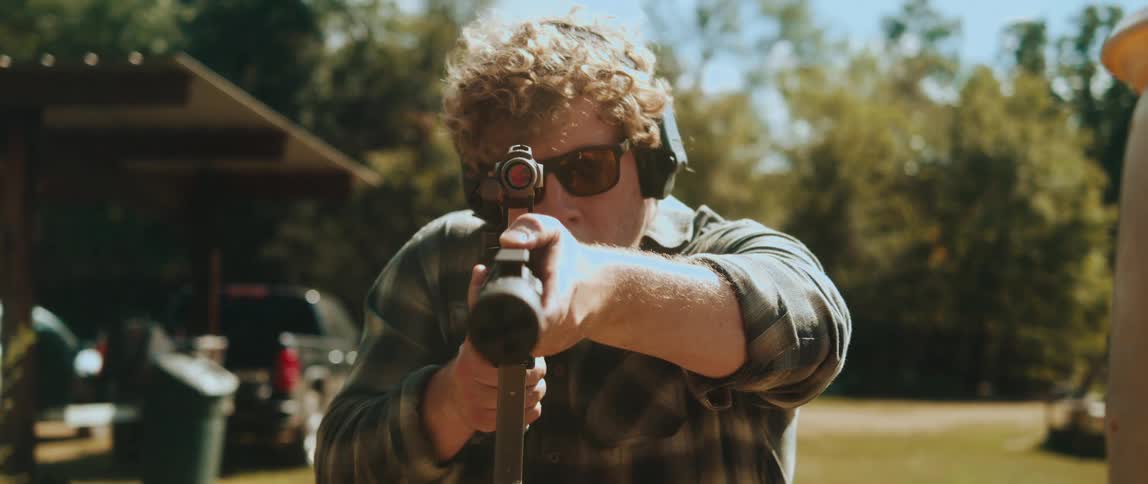 A man with curly hair wearing sunglasses and ear protection aims a rifle directly at the camera the scene takes place outdoors on a sunny day at what appears to be a shooting range