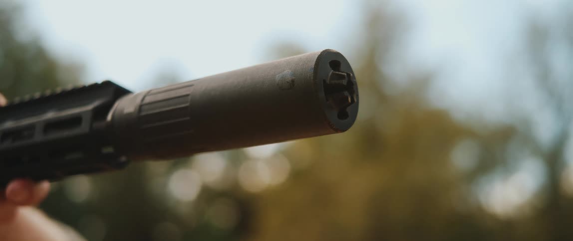 A closeup shot of a black suppressor attached to a rifle barrel as it fires showing the muzzle device vibrating slightly against a blurred background of trees and a bright sky during the day