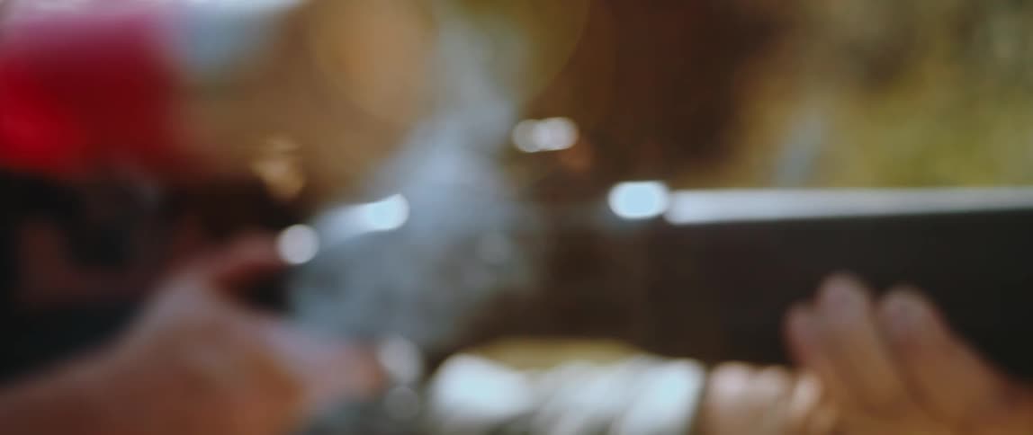 A man fires a shotgun in slow motion causing a red shell to eject from the chamber while smoke trails from the barrel against a blurred outdoor background