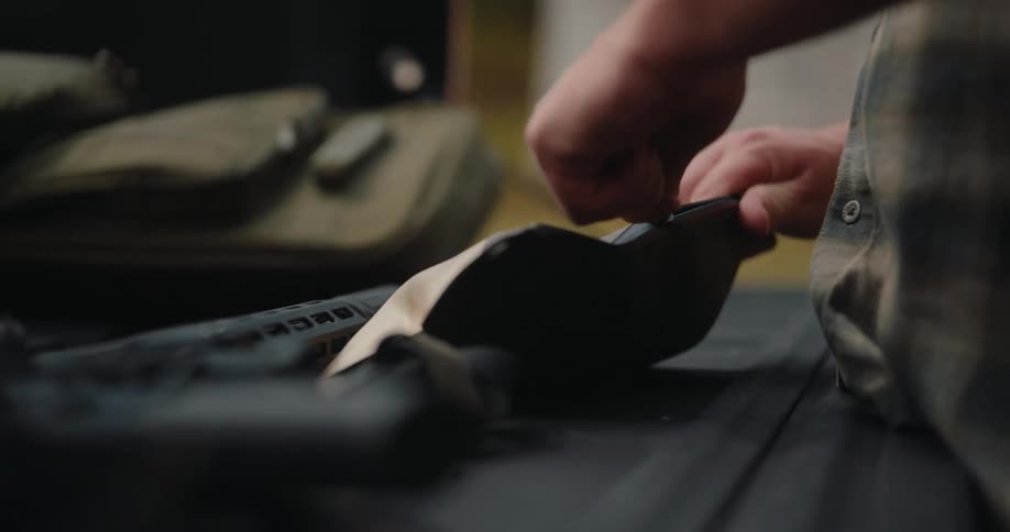 A person is shown working with gear on a flat surface likely a table or tailgate here are two lines describing the scenea persons hands are seen adjusting or securing a piece of black tactical equipment possibly a pouch or holster while a rifle li
