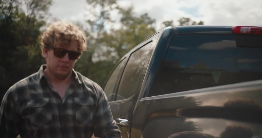 A man wearing sunglasses and a plaid shirt steps out of an open pickup truck door and walks to the tailgate with the vehicles dark exterior reflecting light and trees from the surrounding outdoor setting