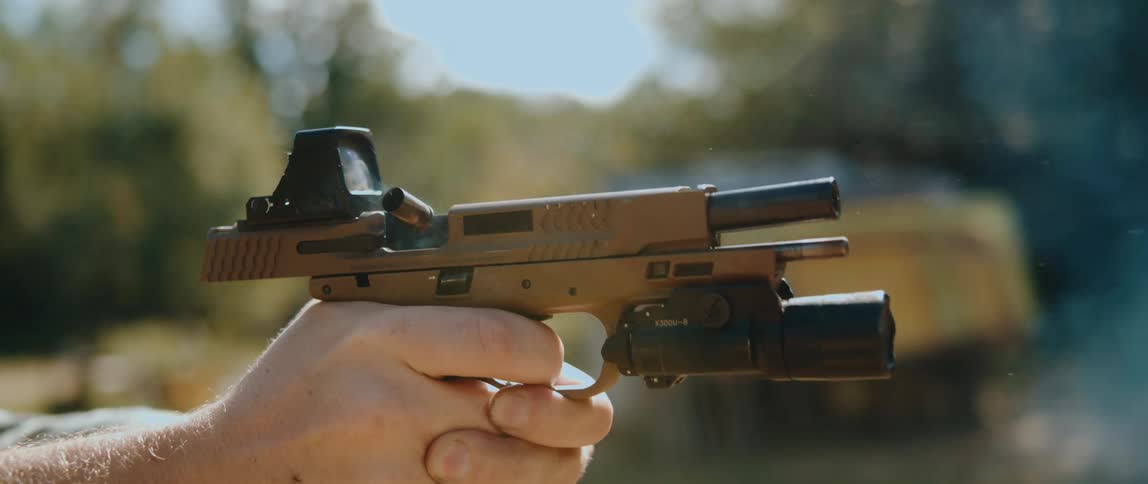 A closeup captures a handgun firing as a spent casing ejects upward from the slide with light smoke visible near the chamber a mounted optic and flashlight attached and a hand firmly gripping the firearm against an outdoor blurred background
