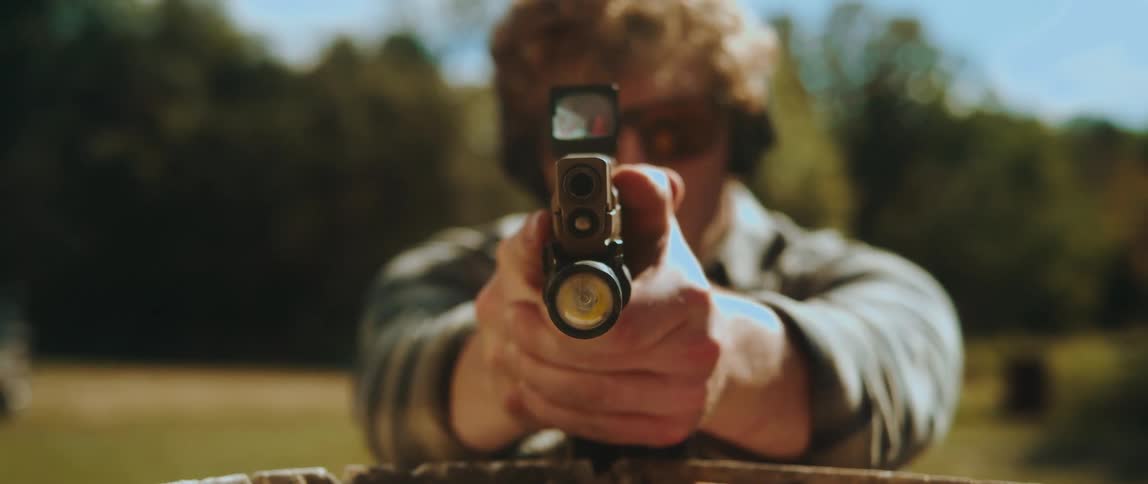 A man in a plaid shirt draws a handgun from his waist and aims it directly at the camera the firearm is equipped with a red dot sight and a tactical light while the shooter wears protective eyewear and earmuffs
