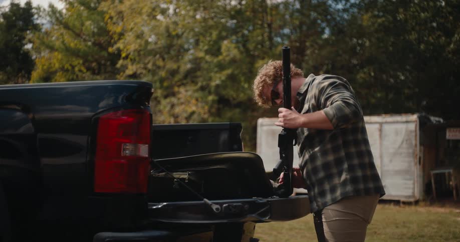 Young adult man wearing sunglasses picking up a rifle and checking its conditions from the trunk of a pickup outdoors during day time