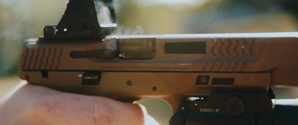A person fires a tan semiautomatic pistol in slow motion showing the slide cycling back a spent shell casing ejecting from the chamber and a puff of smoke rising while the shooter maintains the grip on the weapon