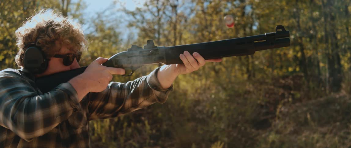 A man with curly hair wearing a plaid shirt and sunglasses fires a shotgun in a forest as a red shell ejects from the chamber