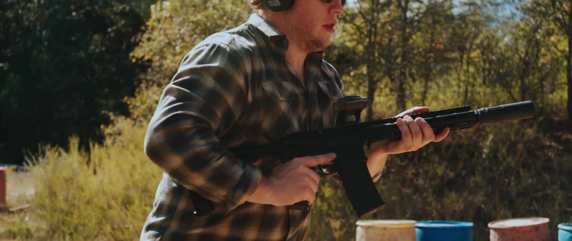 A man in a plaid shirt walks in slow motion while holding an assault rifle aiming forward through a wooded area